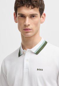 White polo shirt with a green and light blue striped collar, featuring a subtle "BOSS" logo in green on the chest. Smooth fabric texture.