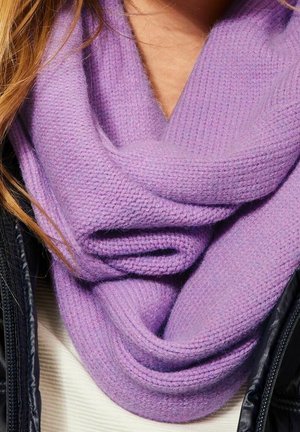 Écharpe infinie en tricot lavande enroulée autour du cou sur un haut blanc et une veste noire, avec des mèches de cheveux blonds visibles.