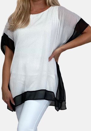 Kobieta ubrana w luźną białą bluzkę w czarne grochy, z czarnymi przezroczystymi rękawami i obszyciem, w połączeniu z białymi spodniami, pozująca z jedną ręką na biodrze.