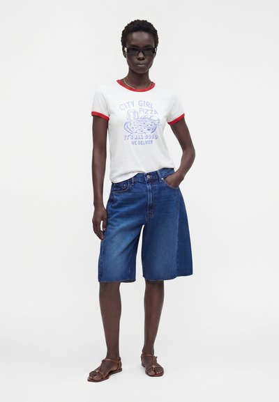 Γυναίκα με γυαλιά ηλίου, λευκό T-shirt με κόκκινη επένδυση και γραφικό με πιτσαρία, άνετα ντένιμ σορτς και καφέ σανδάλια, στέκεται με το ένα χέρι στην τσέπη.