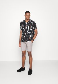 Chemise noire à manches courtes avec des motifs abstraits blancs et orange, associée à un short beige clair et des chaussures noires. Design simple et moderne.