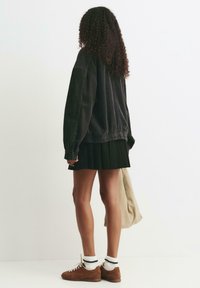 Mörk manchestervägg, plisserad svart kjol, bruna mockaskor, vita strumpor med svarta ränder. Modell som vänder ryggen mot kameran med en beige väska.