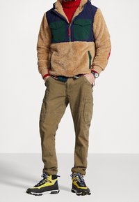 Pullover in fleece con blocchi di colore beige, blu navy e verde; pantaloni cargo marroni con tasche; scarponi da trekking gialli e neri con lacci blu.