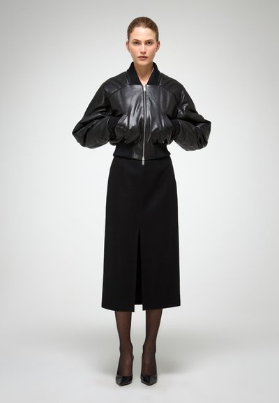 Giacca bomber nera in pelle con maniche oversized, abbinata a una gonna nera aderente con uno spacco frontale. La modella indossa tacchi neri.