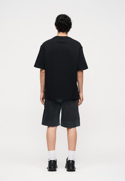 Άτομο με σγουρά μαλλιά στέκεται με την πλάτη γυρισμένη, φοράει ένα μαύρο φαρδύ μπλουζάκι, σκούρα σορτς μέχρι το γόνατο, άσπρες κάλτσες και μαύρα αθλητικά παπούτσια.