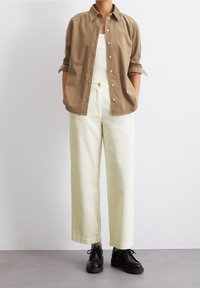 Hellbraunes Button-up-Hemd über einem weißen Tanktop; cremefarbene Cordhosen; schwarze Schnürschuhe; lässige Passform.