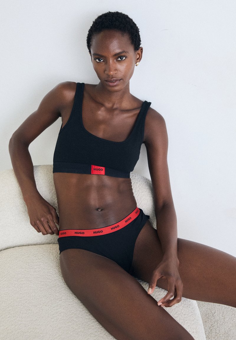 Femme portant un soutien-gorge de sport noir et des sous-vêtements avec un logo Hugo rouge, assise sur une chaise texturée de couleur claire contre un mur uni.