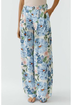 Wijde crèmekleurige broek met grote blauwe bloemenprint, gedragen met een beige top en witte sandalen, hand zichtbaar in de zak.