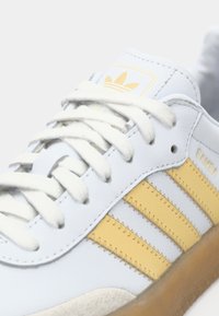 Biały skórzany sneaker z trzema żółtymi paskami, teksturowaną gumową podeszwą oraz beżowym zamszowym noskiem. Posiada płaskie białe sznurówki i logo na języku.