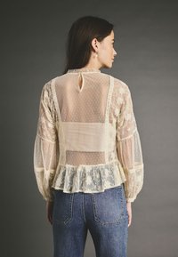 Blusa in pizzo color crema con retro trasparente e tessuto a pois, caratterizzata da un orlo peplum, ricami floreali e apertura a goccia sul collo.