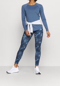 Blaues Langarmoberteil mit rundem Ausschnitt, kombiniert mit dunkelblauen Batik-Leggings. Verfügt über weiße Krawatten-Taille und weiße Sportschuhe.