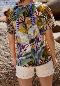 T-shirt à manches courtes avec un motif floral et de feuilles coloré. Fabriqué en tissu léger, il présente une ouverture dans le dos et une coupe décontractée. Associé à un short blanc.