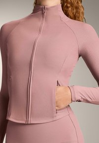 Light pink athletic zip-up jacket with ribbed texture, high collar, and an open pocket on the side. Form-fitting and seamless design.