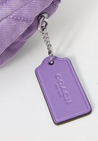 Lila tygpåse med silverdetaljer, med en texturerad lila etikett präglad med "COACH" fäst vid en silverbollkedja.