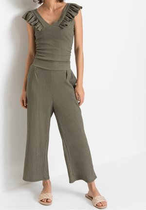 Jumpsuit verde oliva, con scollatura a V arricciata, tessuto testurizzato, design a gamba larga e tasche laterali; abbinato a sandali a infradito intrecciati.