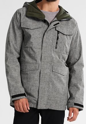 Homme portant une veste à capuche grise avec fermeture éclair et poches à rabat, et des sangles noires aux poignets sur une chemise grise.