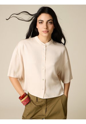 Donna con lunghi capelli scuri sciolti, indossa una camicia beige a maniche corte abbottonata, pantaloni color oliva e bracciali impilati rossi e dorati.