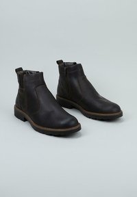 Par de botas de tobillo de cuero marrón oscuro con cremallera lateral y lengüetas, con detalles cosidos y suelas de goma resistentes sobre un fondo gris claro.