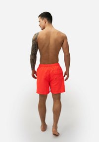 Neon oranje zwemshorts met een elastische tailleband, achterzak en een gladde, lichte stof. Gezichtspunt van achteren, model staat met de rug naar de camera.
