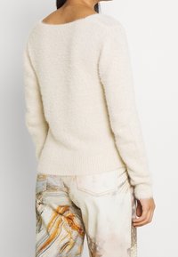 Cremfarbener, flauschiger Strickpullover mit weitem Ausschnitt und gerippten Bündchen, kombiniert mit gemusterten, hellen Jeans.