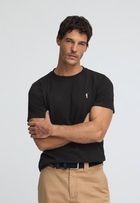 Svart bomulls-t-shirt med en diskret vit logotyp, rund halsringning och korta ärmar, kombinerad med beiga byxor och ett svart bälte.