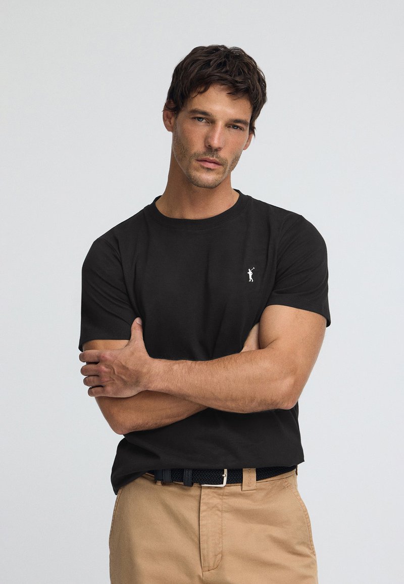 Svart bomulls-t-shirt med en diskret vit logotyp, rund halsringning och korta ärmar, kombinerad med beiga byxor och ett svart bälte.