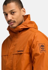 Orange vattentät jacka med huva, utrustad med två bröstfickor, en logopatch på ärmen och kontrasterande svarta sömmar.