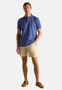 Zila kokvilnas polo krekls ar īsām piedurknēm, apvienots ar pelēkām šortiem un brūnām šņorējamām kurpēm. Krekla krūtīs ir neliela logo.