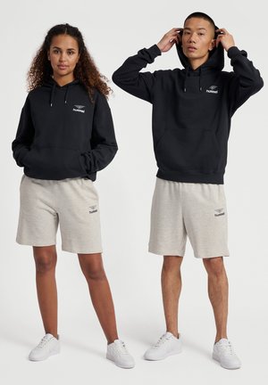 Czarna bluza z kapturem z kieszenią z przodu i logo, w połączeniu z szarymi szortami. Obaj modele mają na sobie białe trampki, co podkreśla swobodny, sportowy styl.