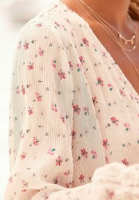 Vaaleanbeige paita, jossa on tekstuurikuvio ja pieniä vaaleanpunaisia ja sinisiä kukkia. Kankaan pinnassa on pehmeää, aaltoilevaa yksityiskohtaa.