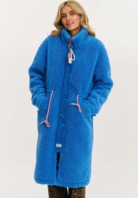 Žena v dlhom, jasnom modrom fleecovom kabáte s ružovými šnúrkami a červeným zipsom, stojaca na jednoduchom pozadí.