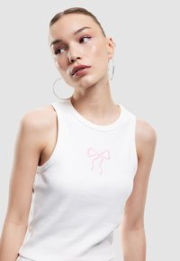 Witte geribde tanktop met een roze geborduurd strikdesign op de borst. De halslijn is rond en de pasvorm is aansluitend.