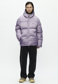 Chaqueta acolchada morada con cuello alto y cierre de cremallera. Presenta secciones acolchadas y es usada sobre pantalones de pana negra y zapatos beiges.