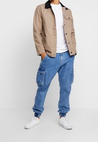 Mies, joka pitää yllä beigeä nappilistallista takkia valkoisen T-paidan, sinisten cargo-farkkujen elastisilla hihoilla ja valkoisten tennareiden kanssa, seisoo kädet taskuissa.