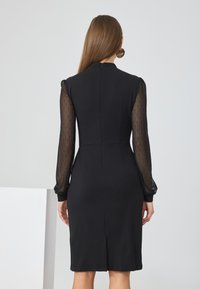 Black dress with long, sheer polka dot sleeves. Features a high neckline, fitted silhouette, and a back slit for ease of movement.