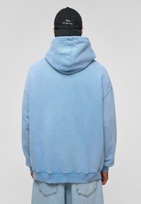 Lichtblauwe oversized hoodie met een gezellige textuur en een grote capuchon, in combinatie met een zwarte pet met witte tekst. Ontspannen pasvorm.