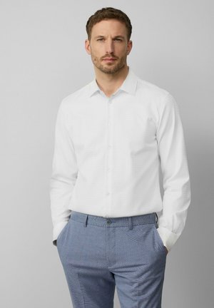 Witte overhemd met lange mouwen en een subtiel gemarmerd patroon, met button-down kraag, gecombineerd met grijze geruite broek. Model staat met handen in de zakken.