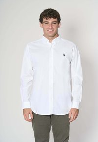 Camicia bianca a maniche lunghe con bottoni, realizzata in cotone, con un piccolo logo di un cavallo blu navy sul lato sinistro del petto, indossata con pantaloni verde oliva.