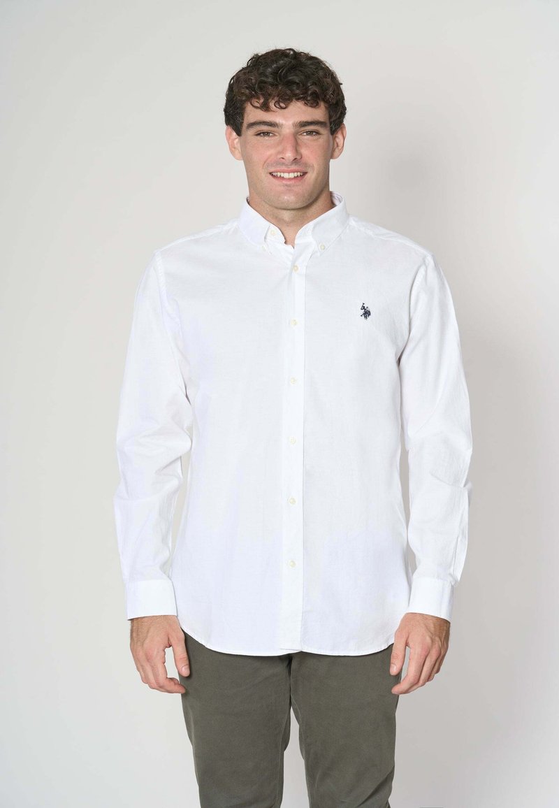 Camicia bianca a maniche lunghe con bottoni, realizzata in cotone, con un piccolo logo di un cavallo blu navy sul lato sinistro del petto, indossata con pantaloni verde oliva.