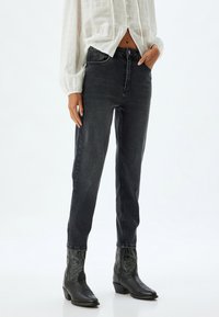 Jeans de denim negro con corte ajustado, que cuentan con un cierre de botón, bolsillos frontales y un leve desgaste. Combinados con una blusa blanca texturizada y botas vaqueras negras.