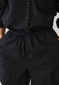 Camisa negra con un bolsillo en el frente y cierre de botones, combinada con pantalones cortos negros con cordón. Ambos están hechos de una tela ligera.