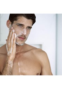 Detergente per il viso applicato sul volto di un uomo, creando una schiuma di bolle. Ha i capelli corti, un viso rasato e la pelle bagnata.