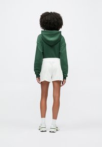 Άτομο με σγουρά μαλλιά που φοράει πράσινο φούτερ με κουκούλα, λευκά σορτς, λευκές κάλτσες και λευκά αθλητικά παπούτσια, στέκεται με την πλάτη στραμμένη σε απλό φόντο.