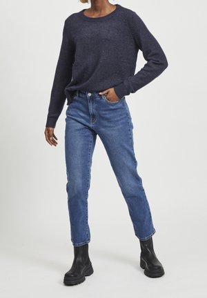Kvinde iført mørkeblå sweater, blå jeans og sorte ankelstøvler, stående med den ene hånd i lommen mod en ensfarvet lys baggrund.