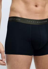 Sorte bomuldsboxershorts med en mørkegrøn talje, prydet med "TOMMY HILFIGER"-logoet i hvid. Ingen mønstre eller yderligere detaljer synlige.