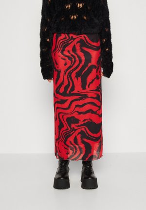 Persona con una falda larga con un patrón abstracto en rojo y negro, suéter negro peludo con agujeros y botas negras de plataforma con cordones.