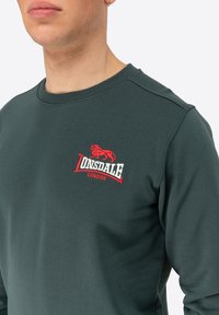 Sötétzöld pulóver kerek nyakkal, bordázott ujjakra, és hímzett vörös oroszlán logóval, alatta "Lonsdale" felirattal.