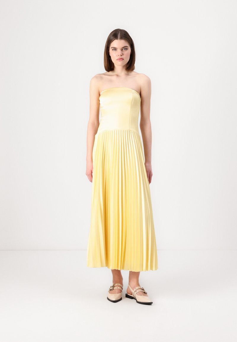 Yellow strapless pleated dress made of smooth fabric, with a fitted bodice and flowing skirt. Paired with two-tone flats.