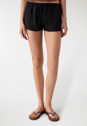 Donna che indossa pantaloncini neri con coulisse e infradito marroni, in piedi davanti a uno sfondo bianco uniforme.