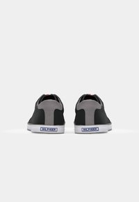 Zapatillas negras con acentos grises, parte superior de textil, suelas de goma blanca y un logo azul en el talón. La vista trasera muestra un cuello acolchado.